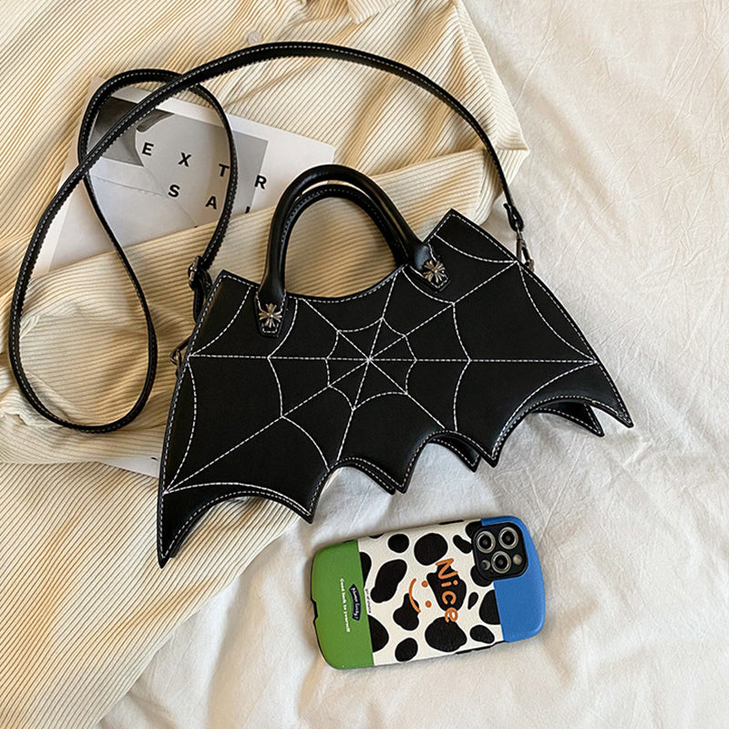 Black bat-wing-shaped bag with its long strap laid on a bed beside a phone.