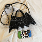 Black bat-wing-shaped bag with its long strap laid on a bed beside a phone.