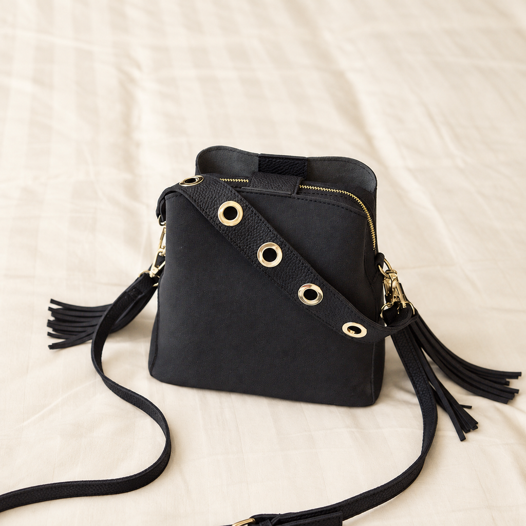Black crossbody bag with gold-tone eyelet strap, zip closure and tassels on an ivory background