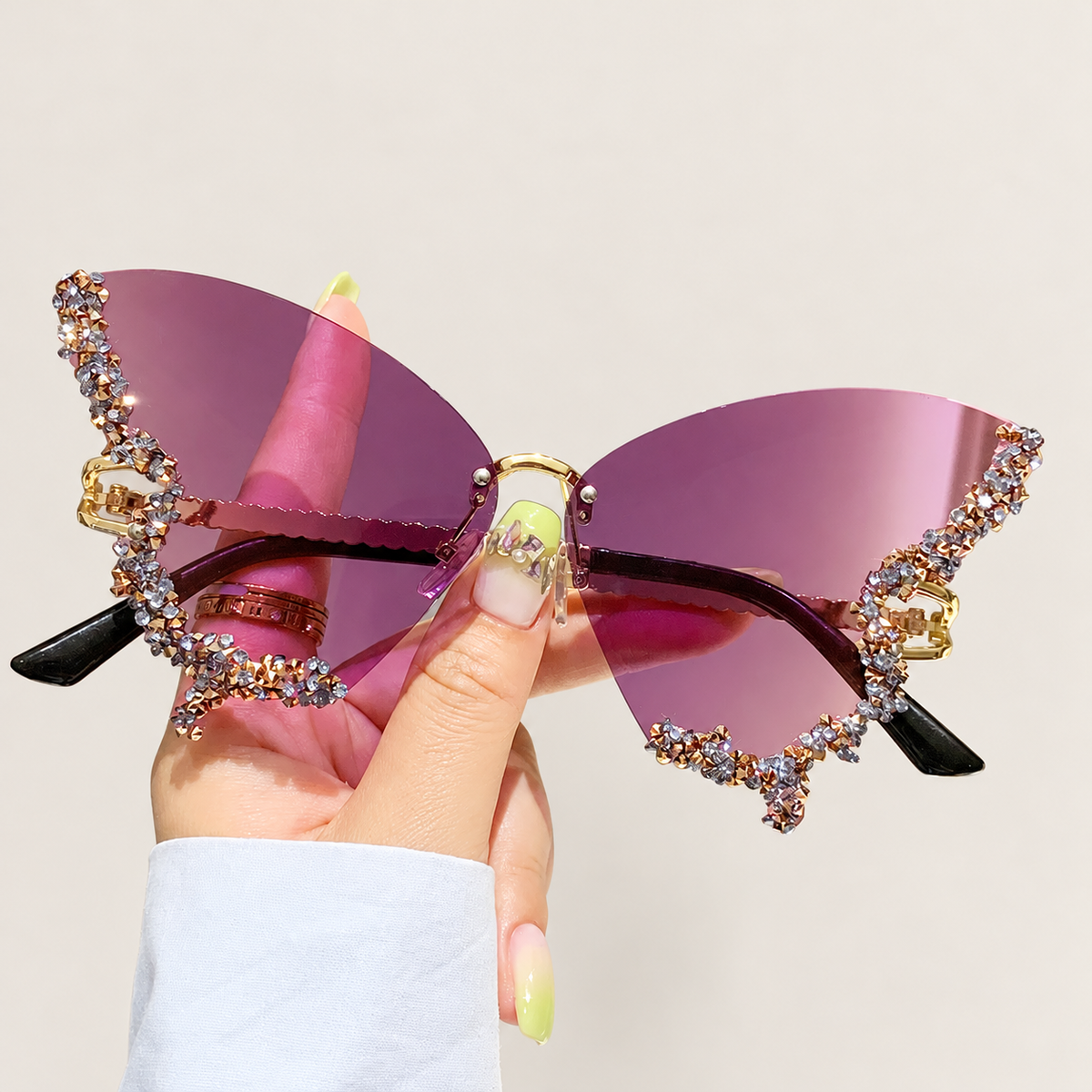 Hand holding purple butterfly-shaped rimless sunglasses with crystal-trimmed edges against a warm ivory background