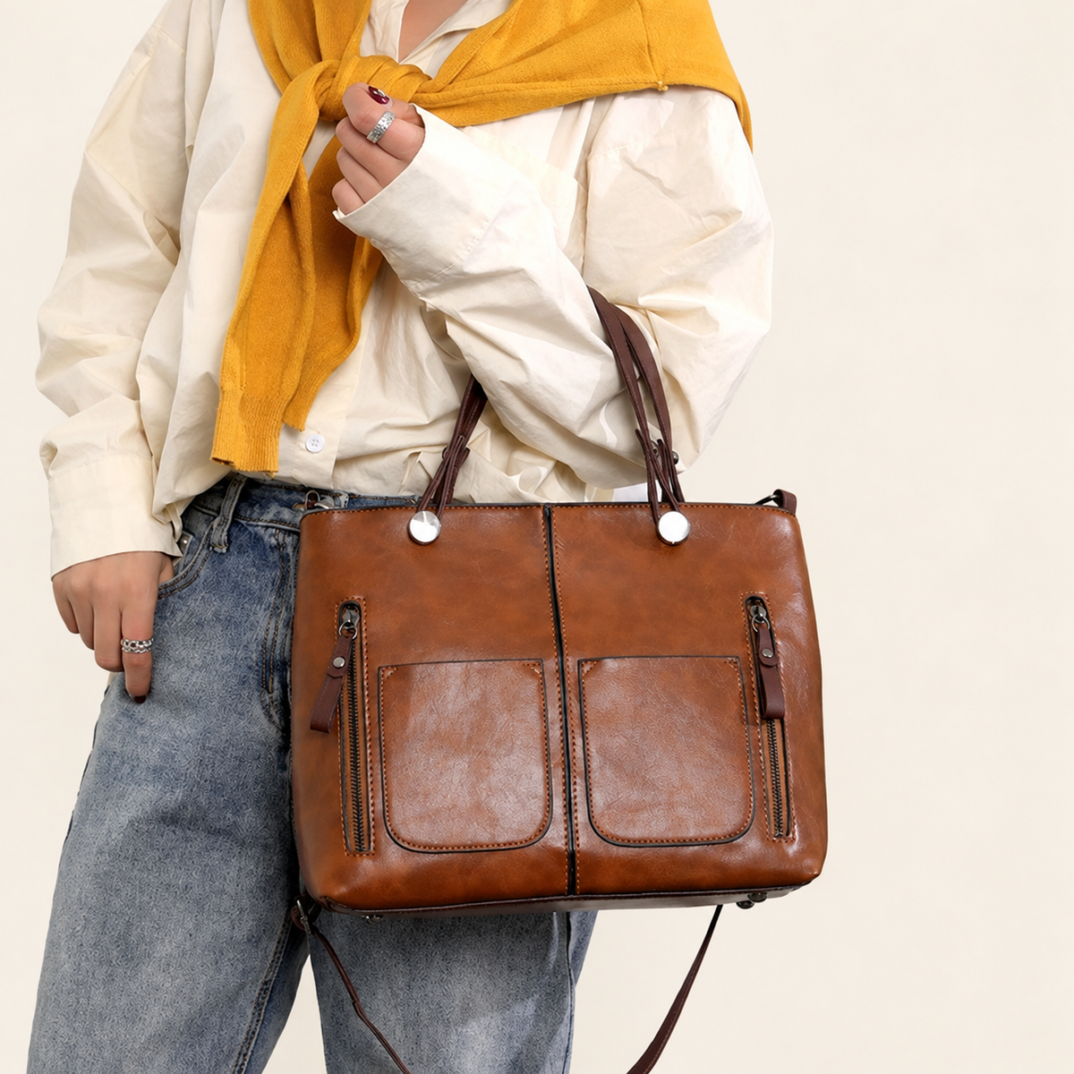 Brown two-handle handbag with front pockets and vertical zippers, carried by a model against ivory