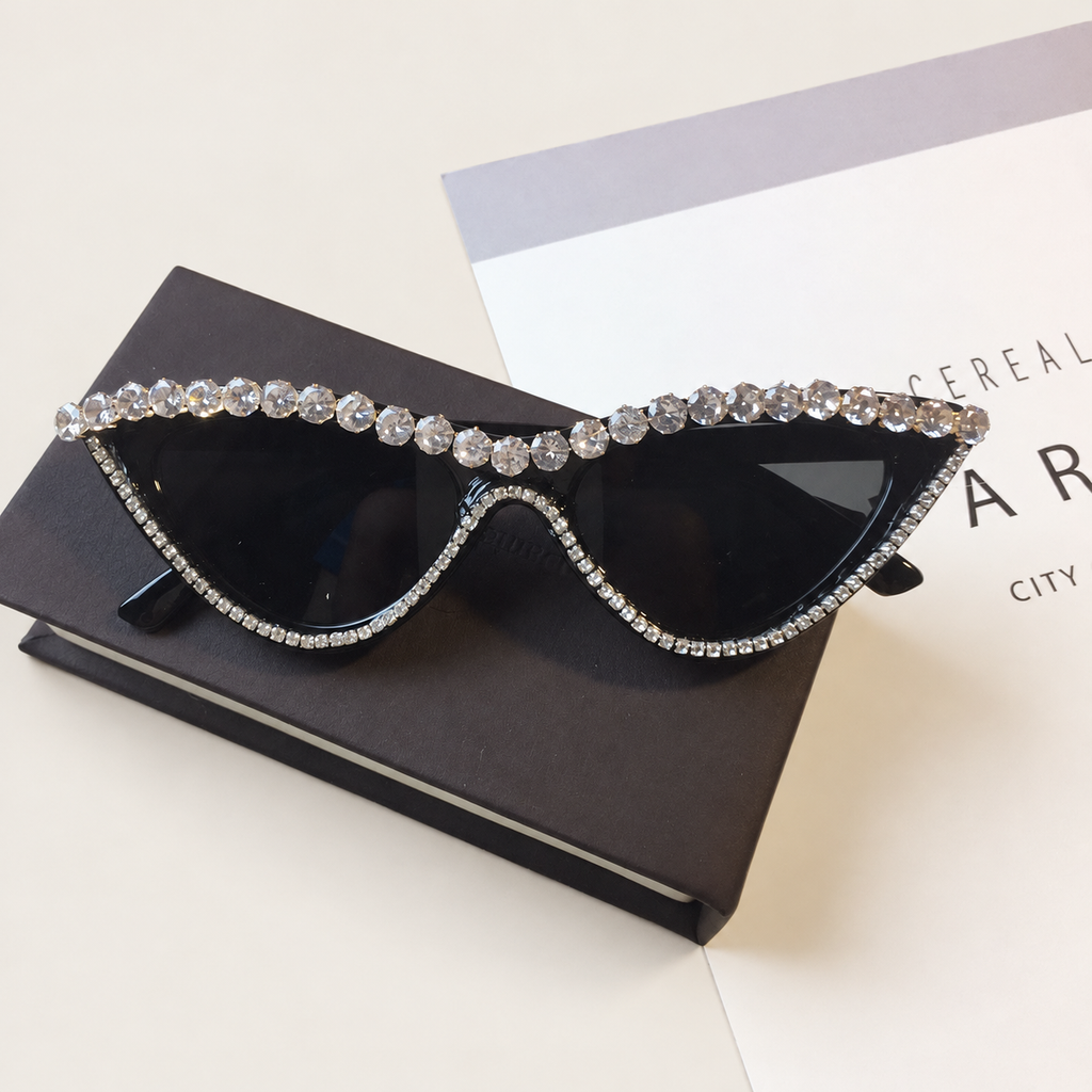 Black rhinestone cat-eye sunglasses resting on a black box beside a printed publication on a warm ivory surface
