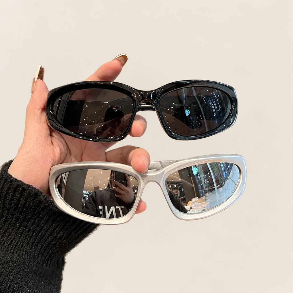 Hand holding black and silver oval sunglasses against a warm ivory studio background