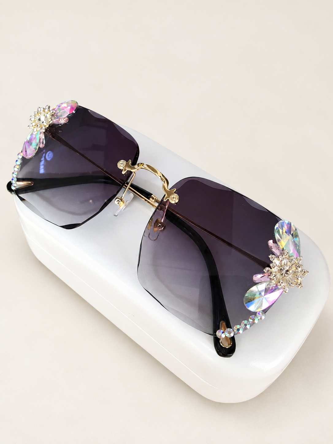 Dark gradient rimless sunglasses with crystal floral embellishments on a white case against a warm ivory background