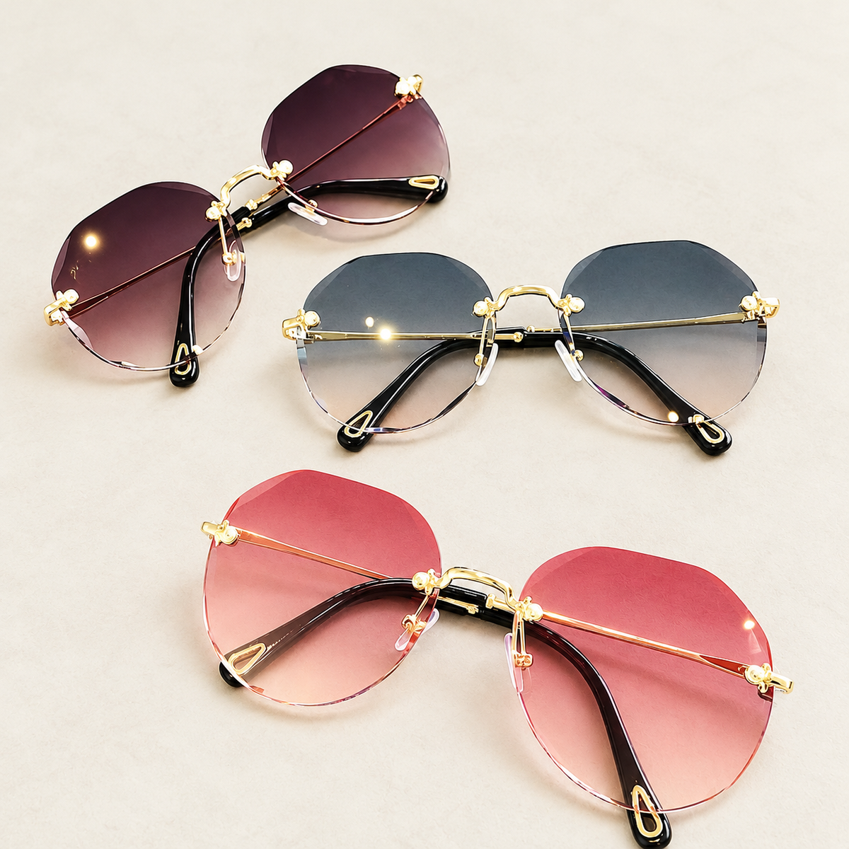 Three pairs of gold-tone rimless polygonal sunglasses in pink and dark gradient lens colors against a warm ivory studio background