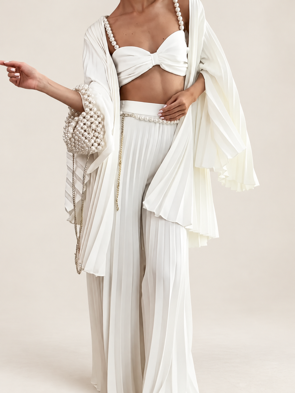 Model wearing a white pearl-strap bandeau, pleated cardigan and wide-leg trousers against a warm ivory studio background