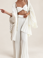 Model wearing a white pearl-strap bandeau, pleated cardigan and wide-leg trousers against a warm ivory studio background