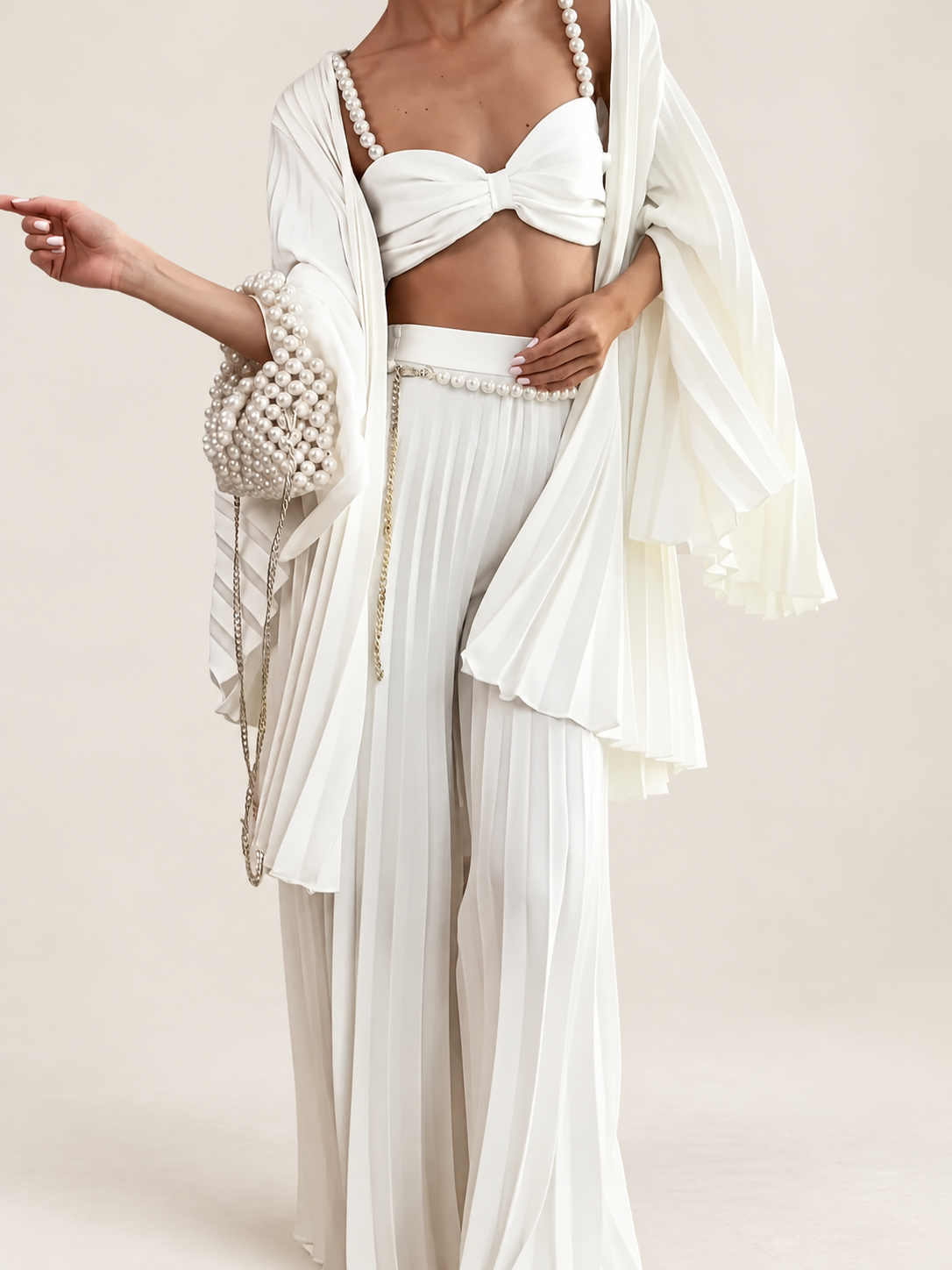 Model wearing a white pearl-strap bandeau, pleated cardigan and wide-leg trousers against a warm ivory studio background