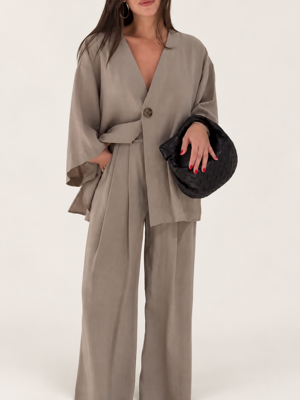 Model wearing a taupe asymmetrical one-button top and wide-leg trousers against a warm ivory studio background