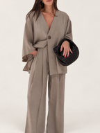 Model wearing a taupe asymmetrical one-button top and wide-leg trousers against a warm ivory studio background