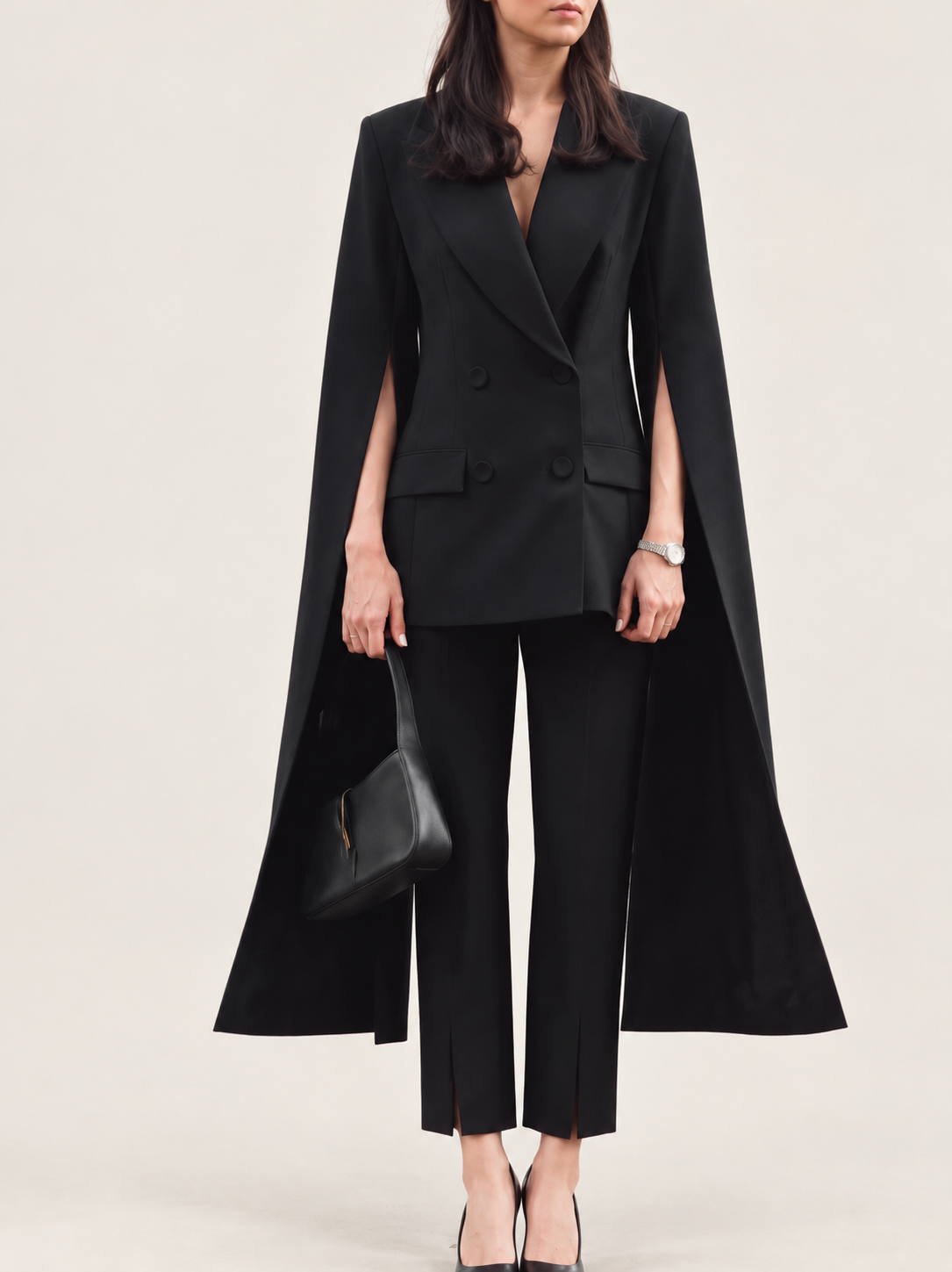 Model wearing a black cape blazer and cropped trousers against a warm ivory studio background