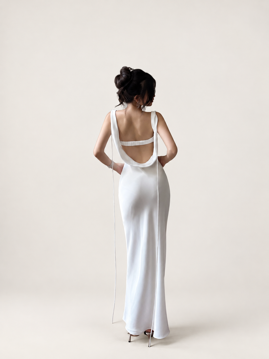 Back view of a white satin maxi dress with an open cowl back and long ties against a warm ivory studio background