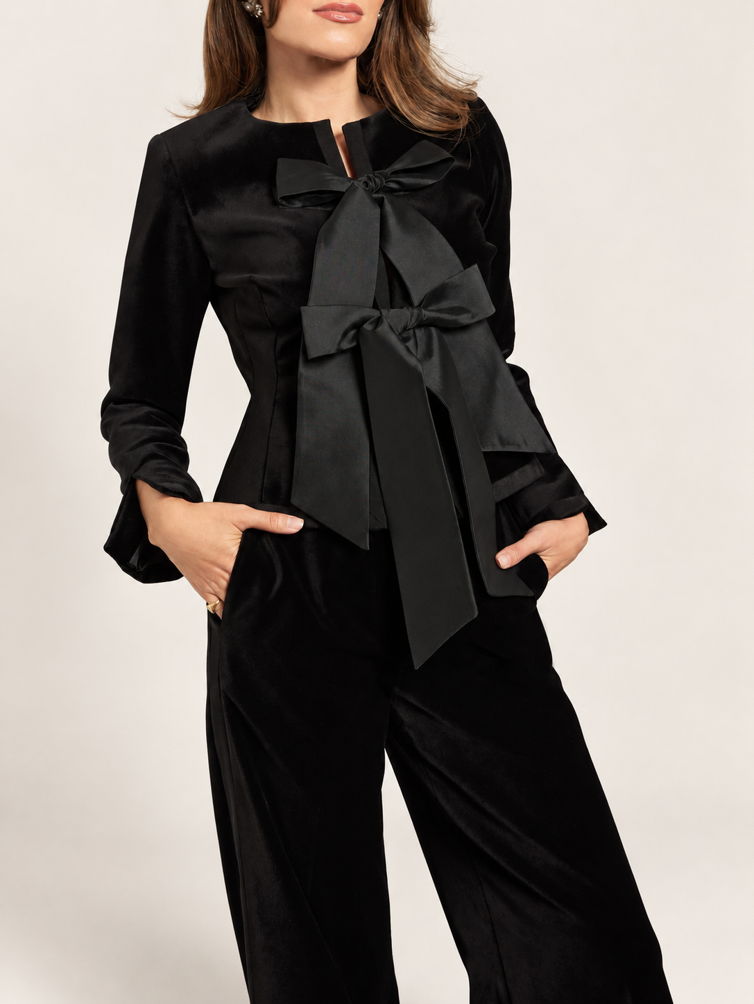 Model wearing a black bow-front jacket and matching trousers against a warm ivory studio background
