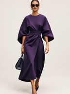 Model wearing a deep purple belted dress with wide sculptural sleeves against a warm ivory studio background
