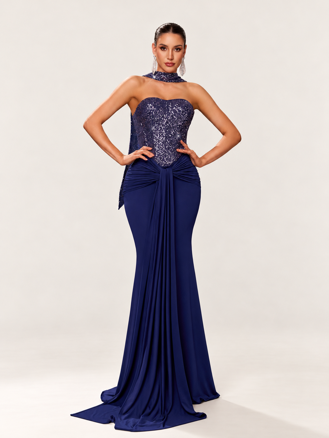 Model wearing a navy sequin-bodice mermaid gown with a matching neck scarf against a warm ivory studio background
