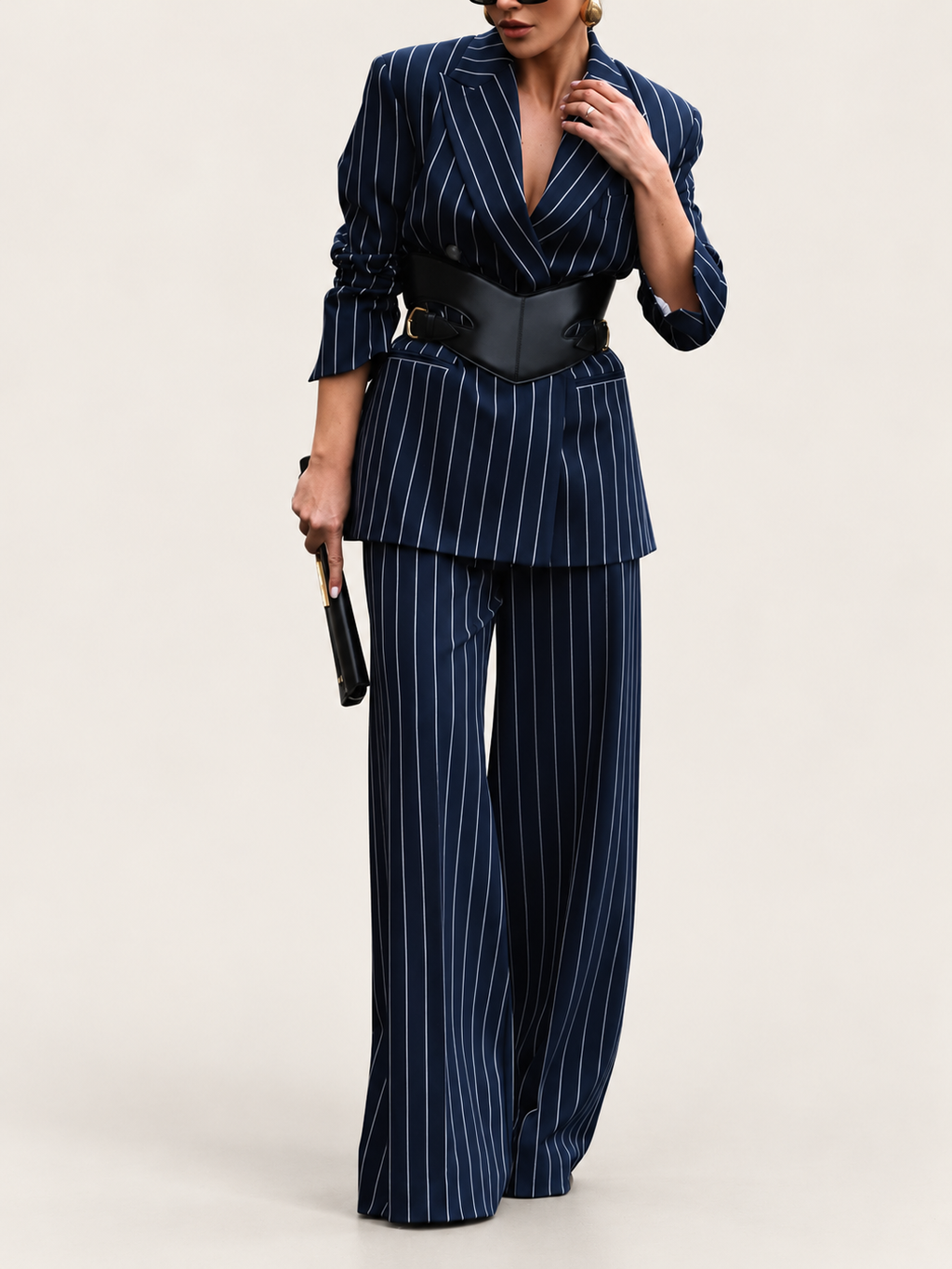 Model wearing a navy pinstripe blazer and wide-leg trousers with a black waist belt against a warm ivory studio background