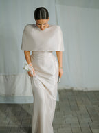 Pale cape-overlay fitted dress worn by a model in front of a neutral curtain