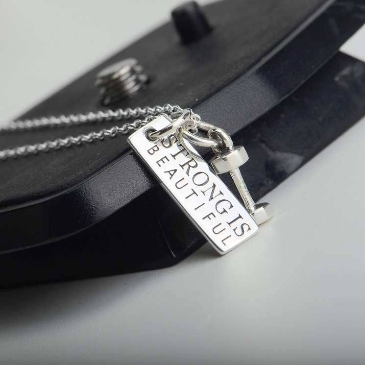 Silver-coloured necklace with a dumbbell charm and a rectangular "STRONG IS BEAUTIFUL" pendant, displayed on a black stand.