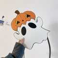 Angled front view of the white ghost-shaped bag with an orange pumpkin hat and black strap held in one hand.