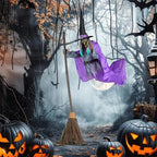 Purple-cloaked witch decoration holding a vertical broom in a Halloween forest display, with a black pointed hat and grey costume.