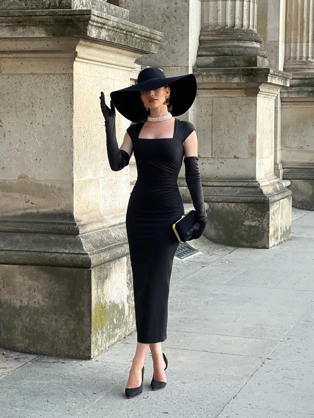 Model wearing a black square-neck midi dress with long gloves; hat, jewellery, clutch and shoes are styling accessories