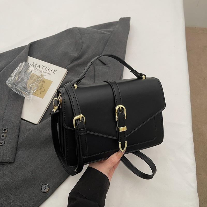 Black handbag with a top handle, shoulder strap and gold-tone clasp, shown held in a hand