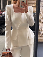 Front view of a white square-neck blazer with covered buttons and a layered hem; trousers and bag are styling accessories