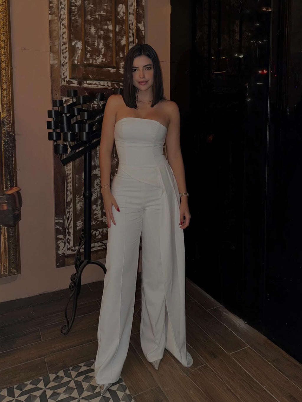 White strapless top and wide-leg trousers worn indoors, showing the long asymmetric side drape.