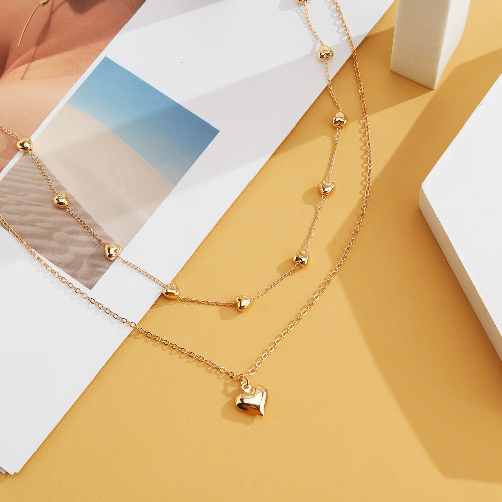 Gold-tone double-layer heart necklace displayed over a white block beside printed pages on a yellow surface.